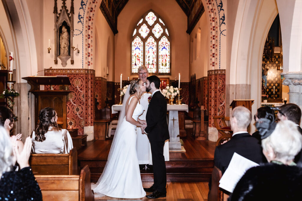 A bride and groom kiss at the altar during their wedding ceremony in a church, with guests seated and a priest standing behind them. The stained glass windows and ornate décor are visible in the background.