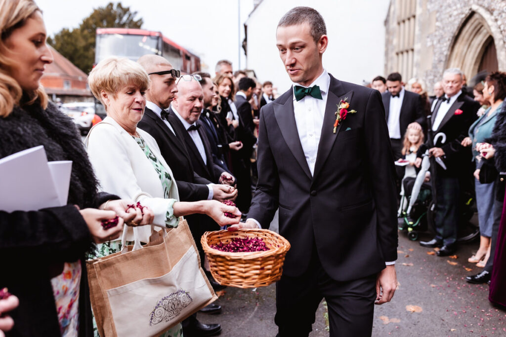 A man in a tuxedo holds a basket of rose petals as wedding guests in formal attire gather outside, reaching in to take petals. A red double-decker bus is visible in the background.