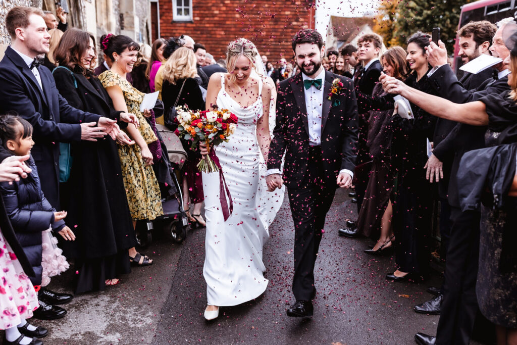 A bride and groom walk hand in hand down an outdoor path, smiling as guests celebrate by throwing confetti. The bride holds a colorful bouquet, and people on both sides cheer, clap, and take photos.