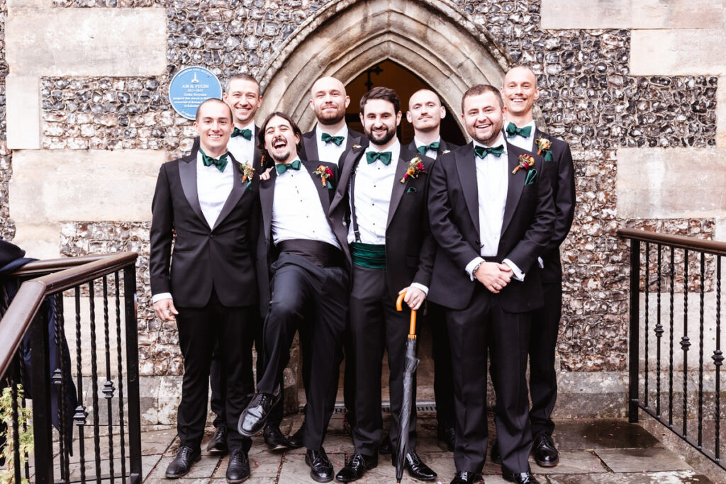 Eight men in tuxedos with green bow ties and cummerbunds stand smiling outside a stone church entrance. One man playfully lifts his leg, while another holds a cane. All look happy and celebratory.