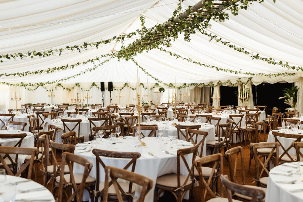 A large, elegant tent is decorated for an event with white draped ceilings, greenery garlands, round tables covered in white cloths, and wooden chairs arranged neatly around each table.
