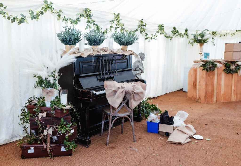 A black upright piano decorated with foliage, bouquets, and burlap bows sits in a tent next to stacked suitcases and a rustic chair. Plants and draped garlands adorn the white tent interior.