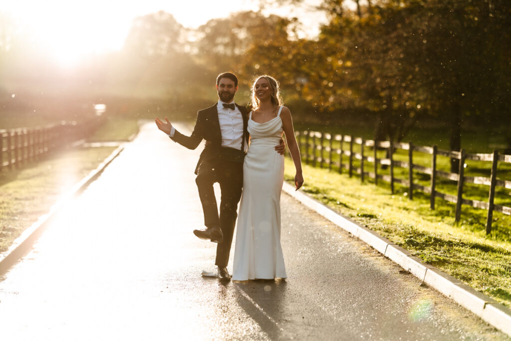 A newly married couple walks joyfully down a sunlit country road. The groom wears a tuxedo, the bride wears a white dress, and they smile, holding each other as golden sunlight shines around them.