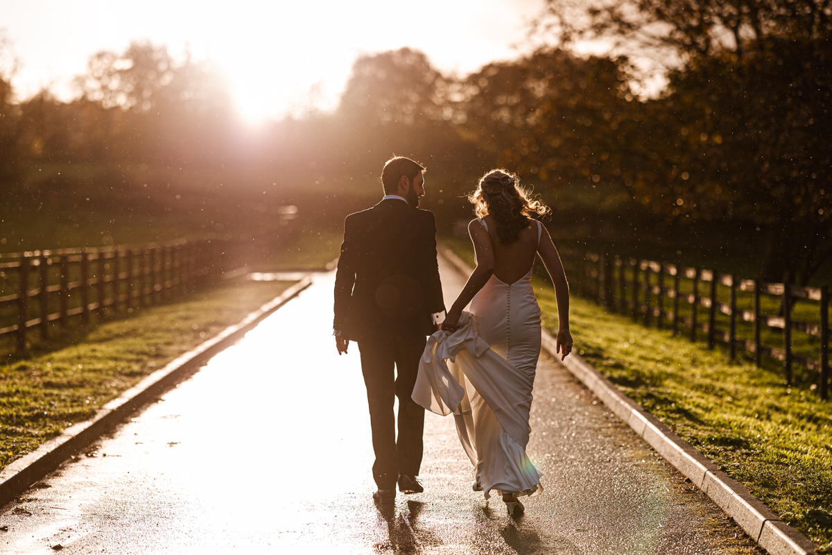 A bride and groom walk hand in hand down a sunlit path, with the glowing sunset behind them and green fields on either side. The bride holds up her dress as they walk together.