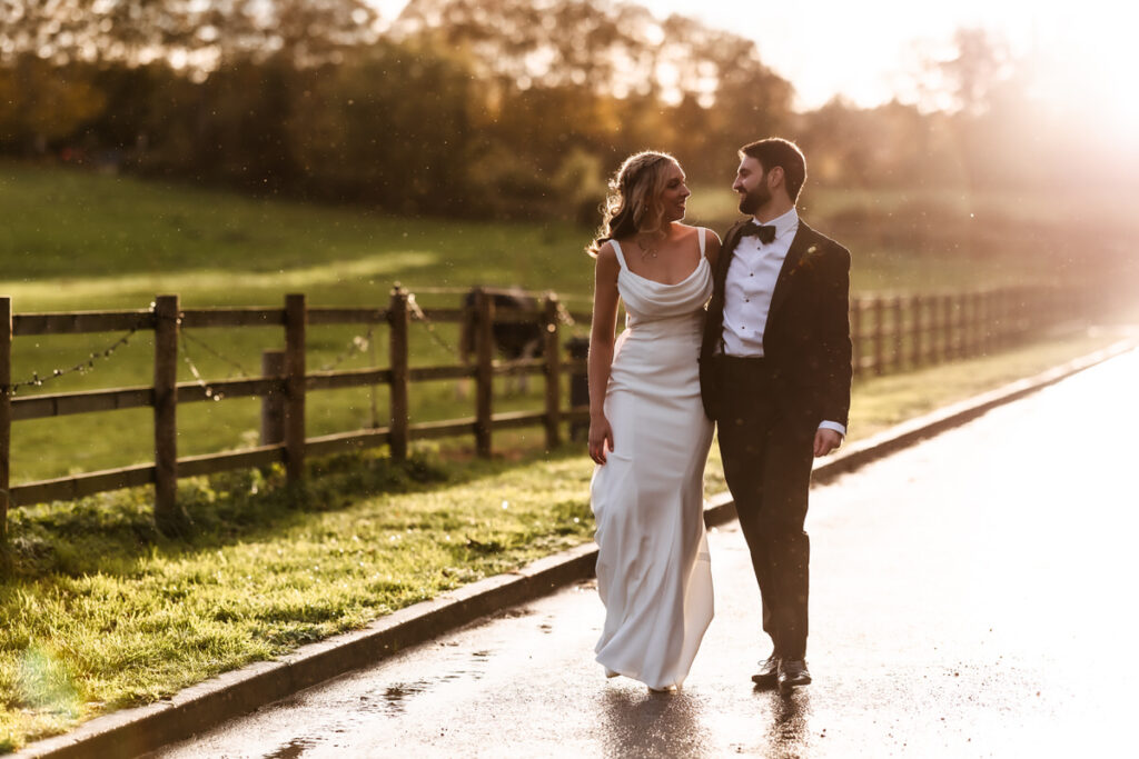 A bride in a white dress and a groom in a black tuxedo walk arm in arm on a sunlit path next to a field, smiling at each other, with trees and a wooden fence in the background.