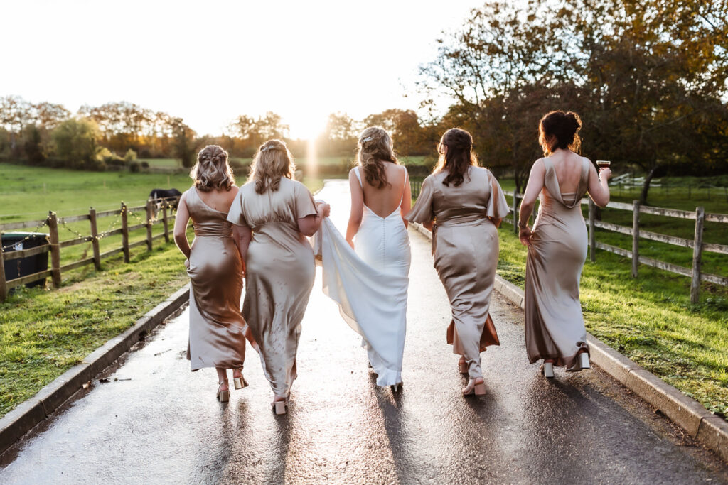 Five women in formal dresses, one in white and four in champagne tones, walk away on a country lane at sunset, with fields and trees in the background.