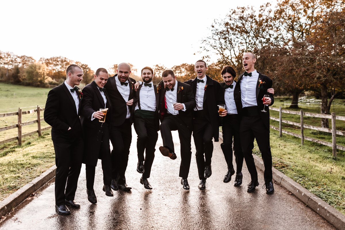 Eight men in tuxedos walk arm-in-arm outdoors on a wet path, smiling and laughing. Some hold drinks, and a few are kicking their legs up. Trees and a wooden fence line the scene on a bright day.
