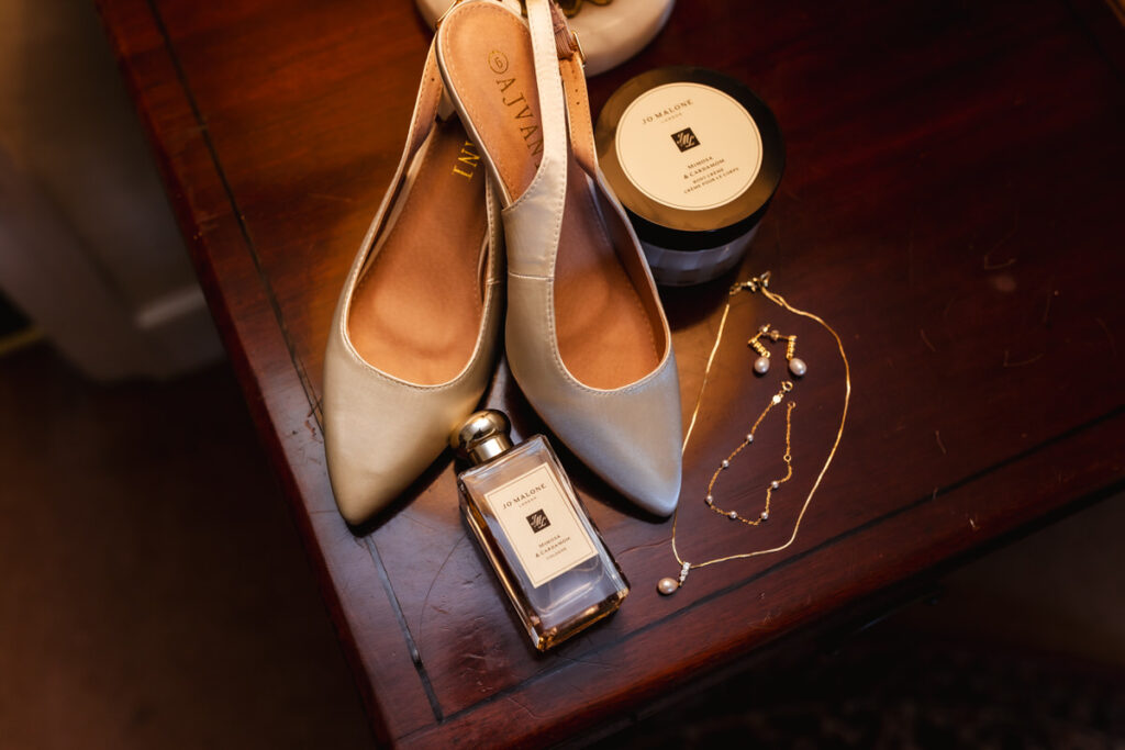 A pair of ivory pointed-toe shoes, a bottle and a jar of Jo Malone perfume, a delicate gold necklace with a pearl, and matching pearl earrings arranged on a wooden table.