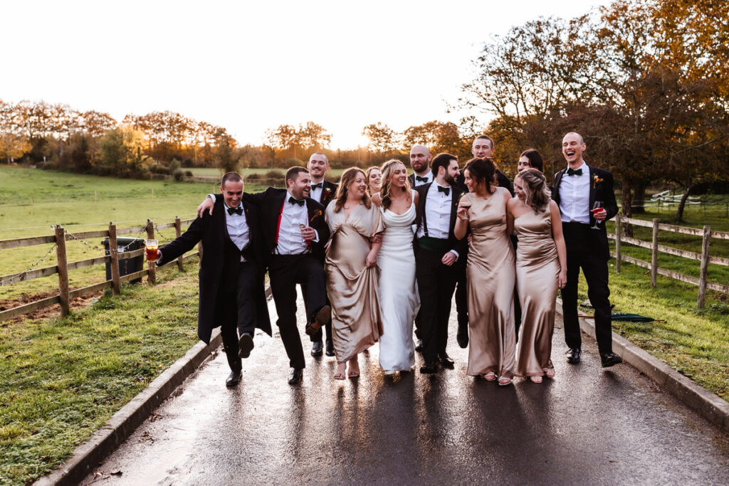 A wedding party walks arm-in-arm on a rural road at sunset, smiling and laughing. The group includes people in formal black suits and women in gold dresses, with green fields and trees in the background.