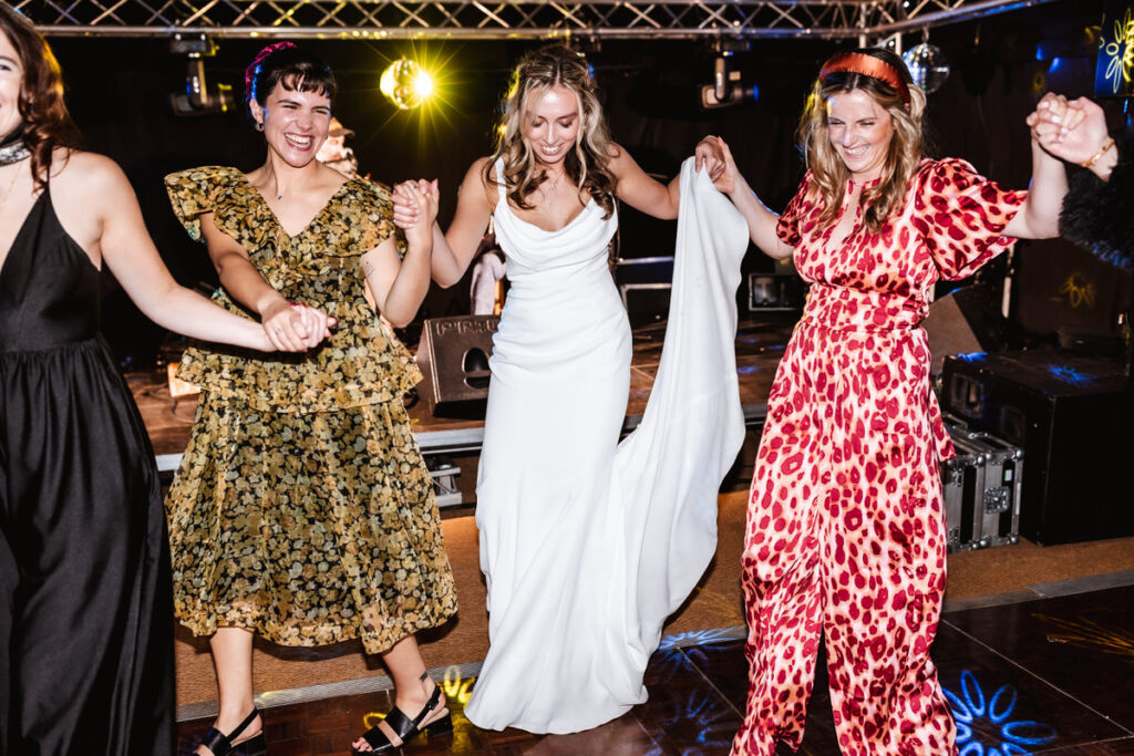 Three women in festive dresses, including one in a white gown, hold hands and dance joyfully at a party or celebration under bright lights.