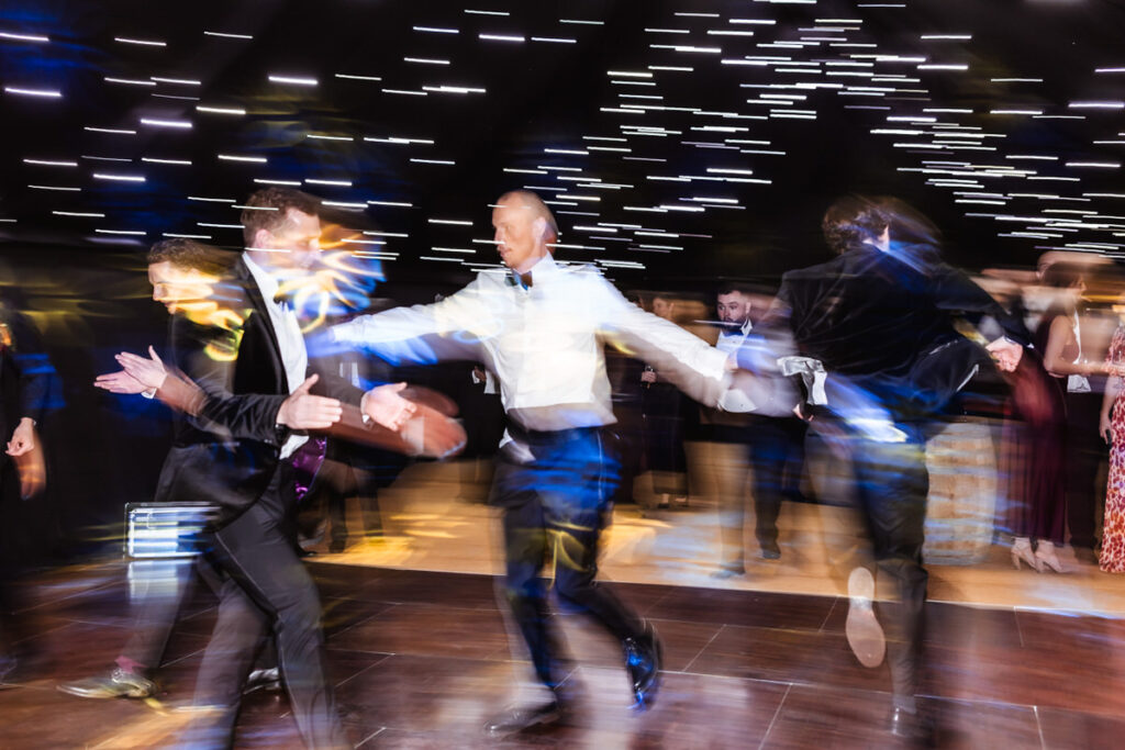 Men in formal wear dance energetically under string lights, creating a motion blur effect. The scene is lively, with guests in the background watching the movement on the dance floor.