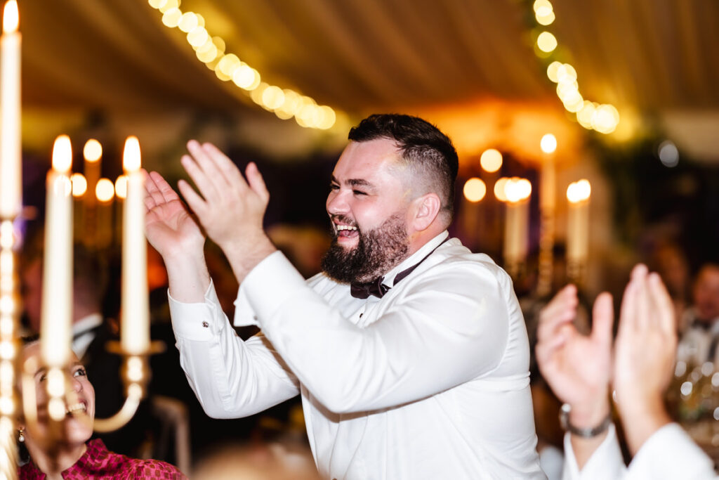 A man in formal attire smiles and claps enthusiastically at an indoor event with warm lighting and blurred candlelights in the background.