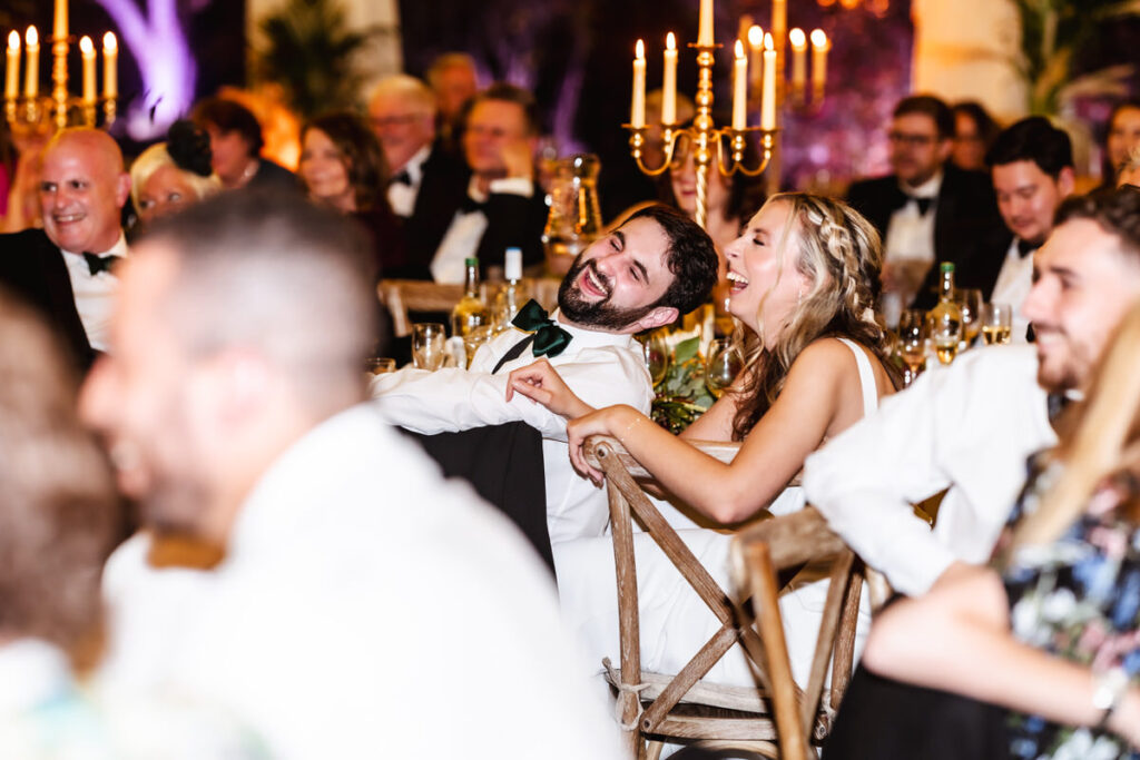 A bride and groom sit together at a reception table, laughing joyfully among well-dressed guests. Elegant candelabras and warm lighting create a festive, celebratory atmosphere.