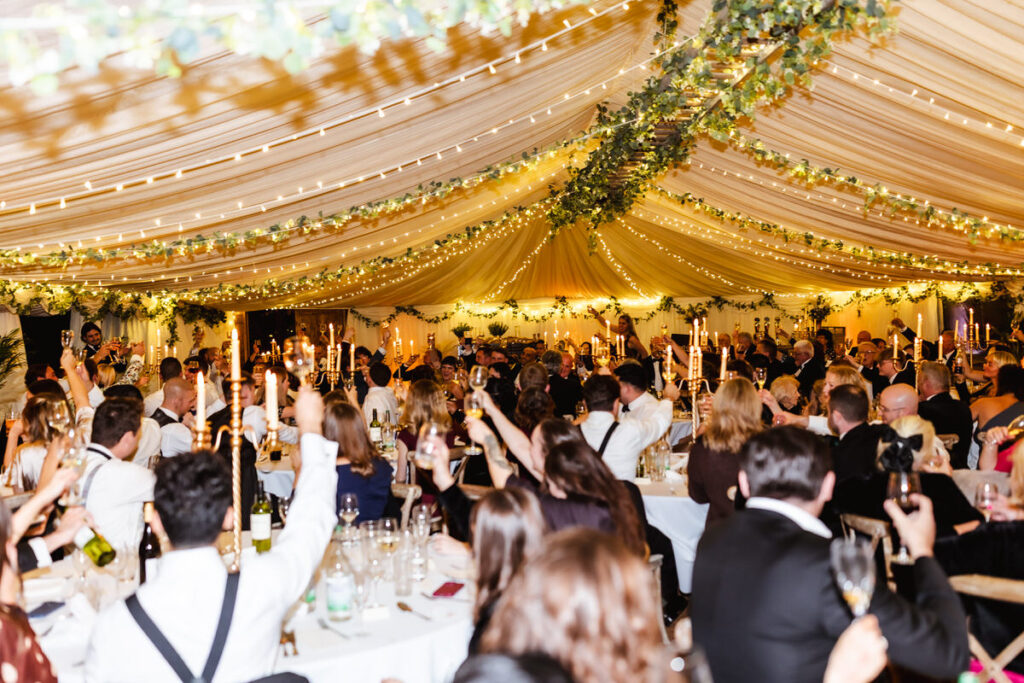 A lively wedding reception under a decorated marquee, with guests seated at long tables, raising glasses in a toast. The ceiling is draped with fabric, greenery, and string lights, creating a festive atmosphere.