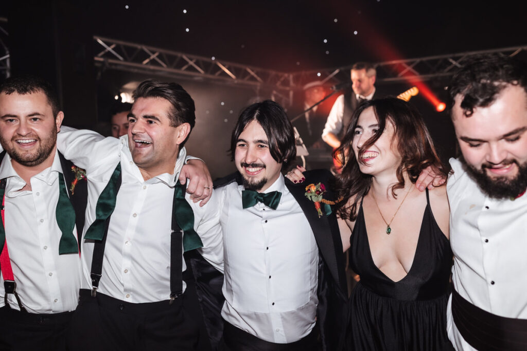 A group of four men and one woman, dressed formally, stand close together with arms around each other, smiling and celebrating at an indoor event with stage lights in the background.