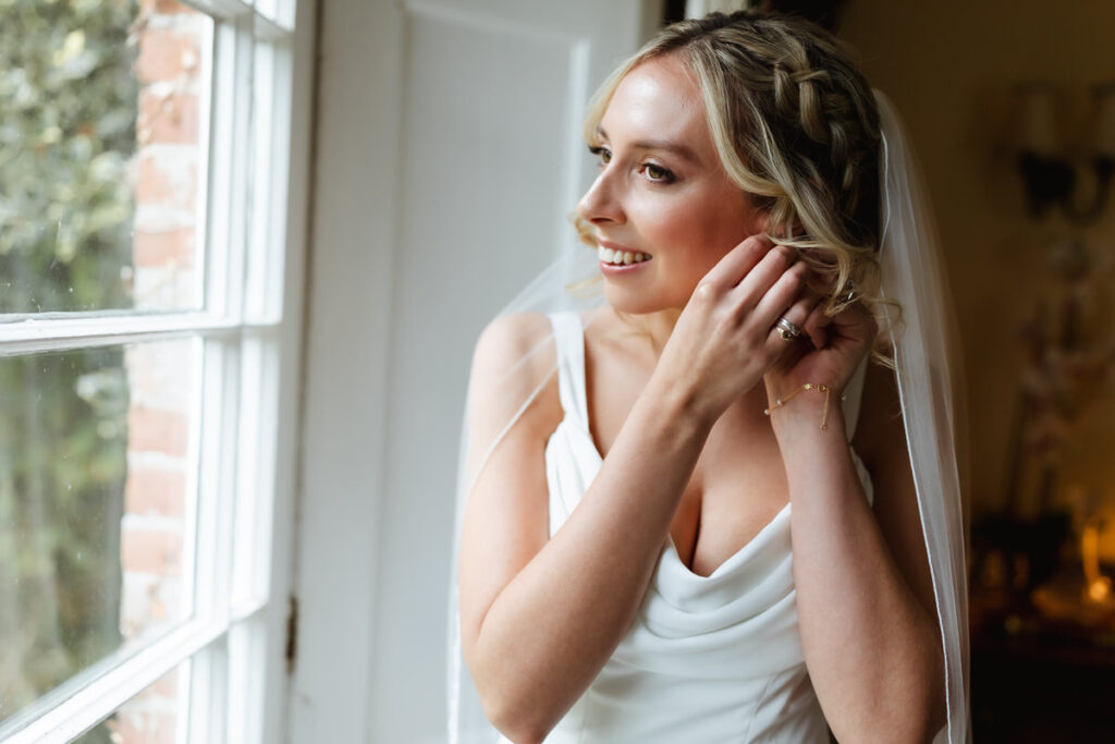 A bride in a white dress and veil smiles as she puts on earrings, standing by a window with soft daylight illuminating her face.