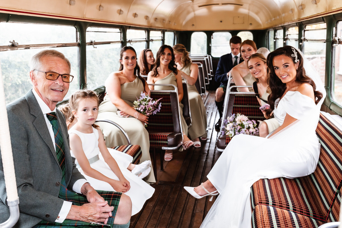 A bride in a white dress sits smiling on a vintage bus with bridesmaids, a flower girl, and an older man in a suit. The group looks cheerful, surrounded by bouquets and patterned bus seats.