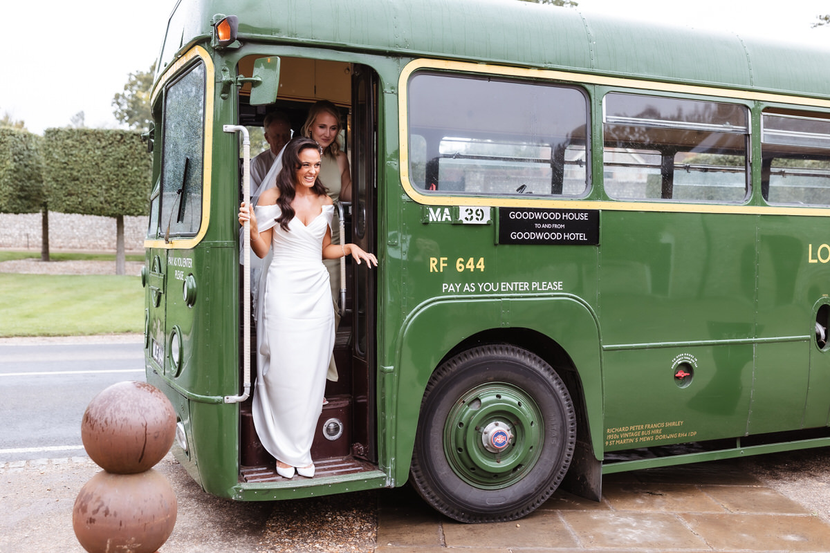 A smiling bride in a white dress steps off a vintage green bus, with another woman in white behind her. The bus has signs for Goodwood House and Goodwood Hotel and is parked near a grassy area.