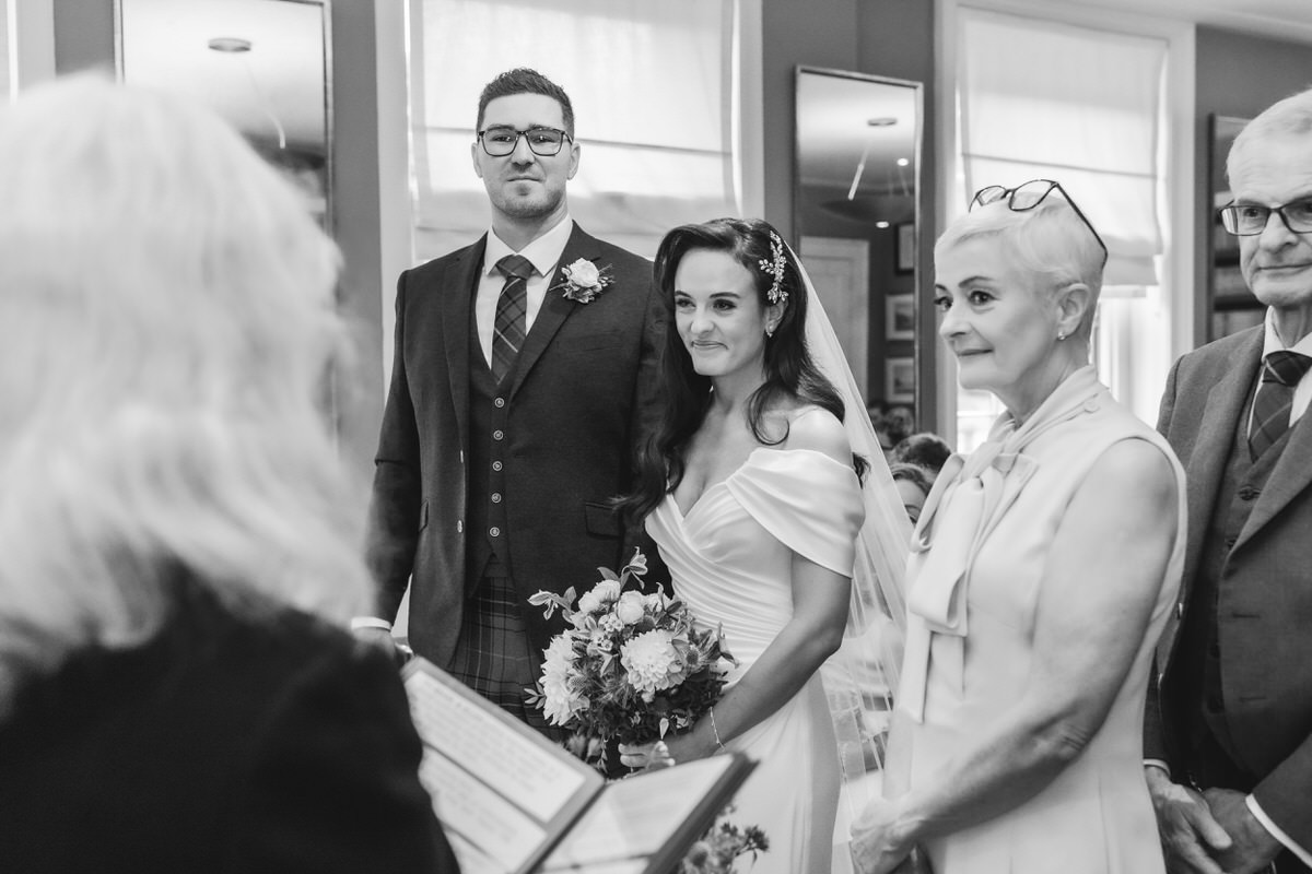 A bride and groom stand together during a wedding ceremony, facing an officiant. The bride holds a bouquet and smiles, with two older guests standing beside her. The scene takes place indoors in a bright room.