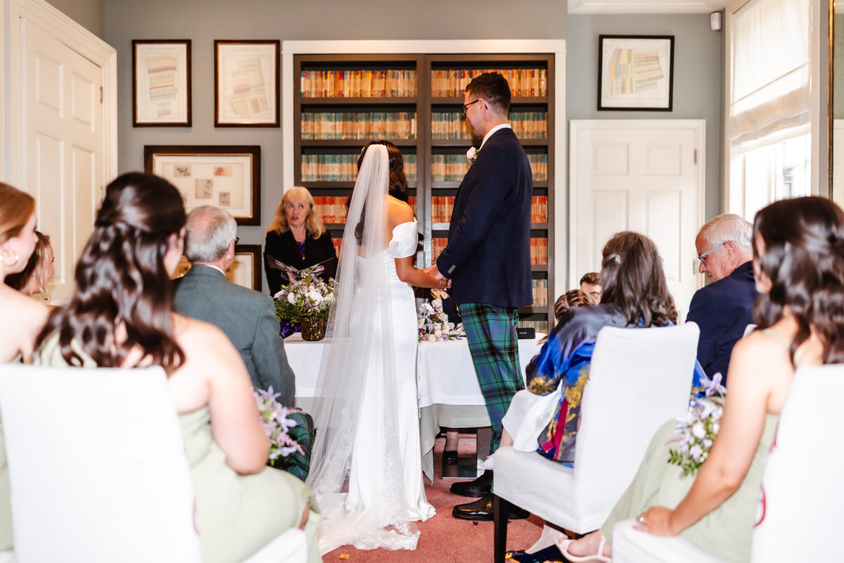 A bride and groom stand facing an officiant during an indoor wedding ceremony, with seated guests watching. The bride wears a white dress and veil; the groom wears a suit with a tartan pattern. Shelves of books line the wall behind them.