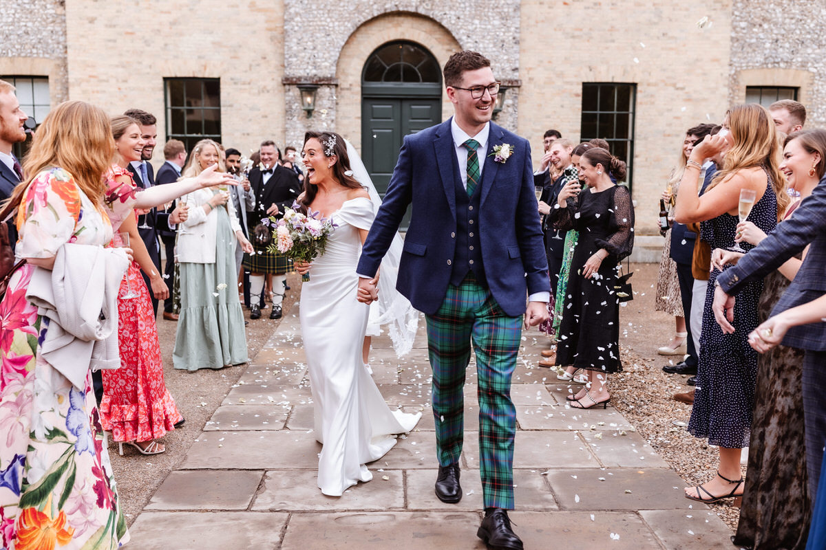 A bride and groom walk hand in hand outside, smiling as guests in colorful outfits toss confetti in celebration. The groom wears a blue jacket and plaid pants; the bride is in a white gown holding a bouquet.