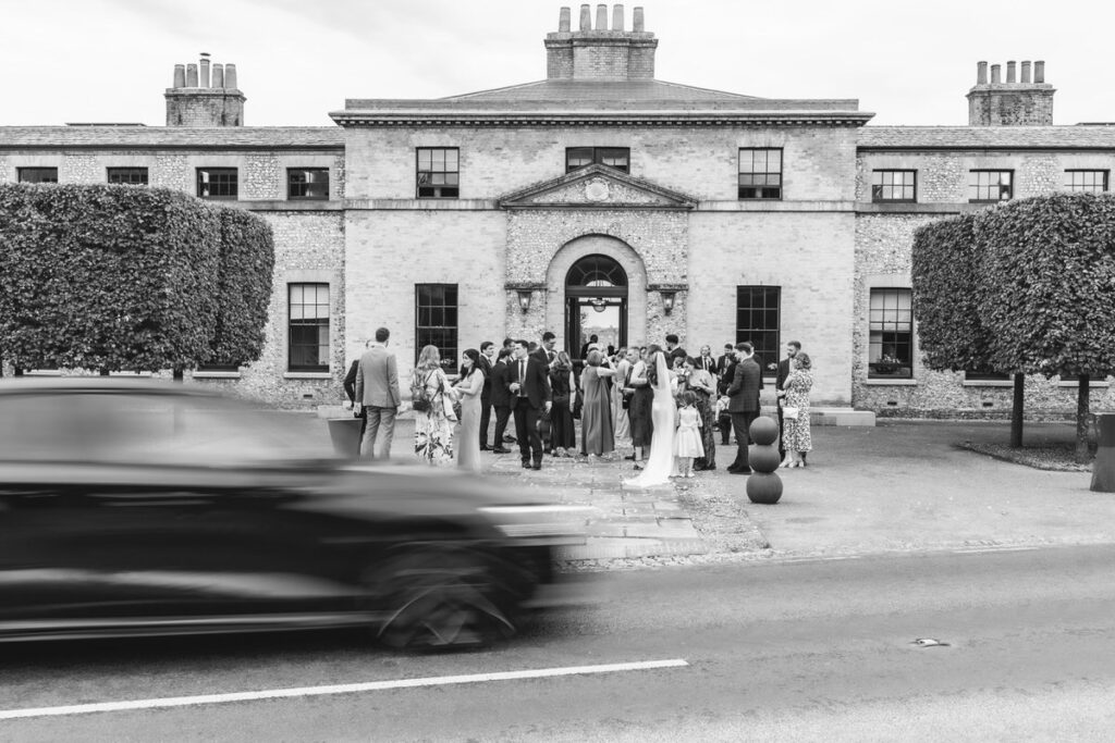 A group of people in formal attire gather outside a large, elegant brick building with trimmed trees. A blurred black car passes by in the foreground on the road. The image is in black and white.