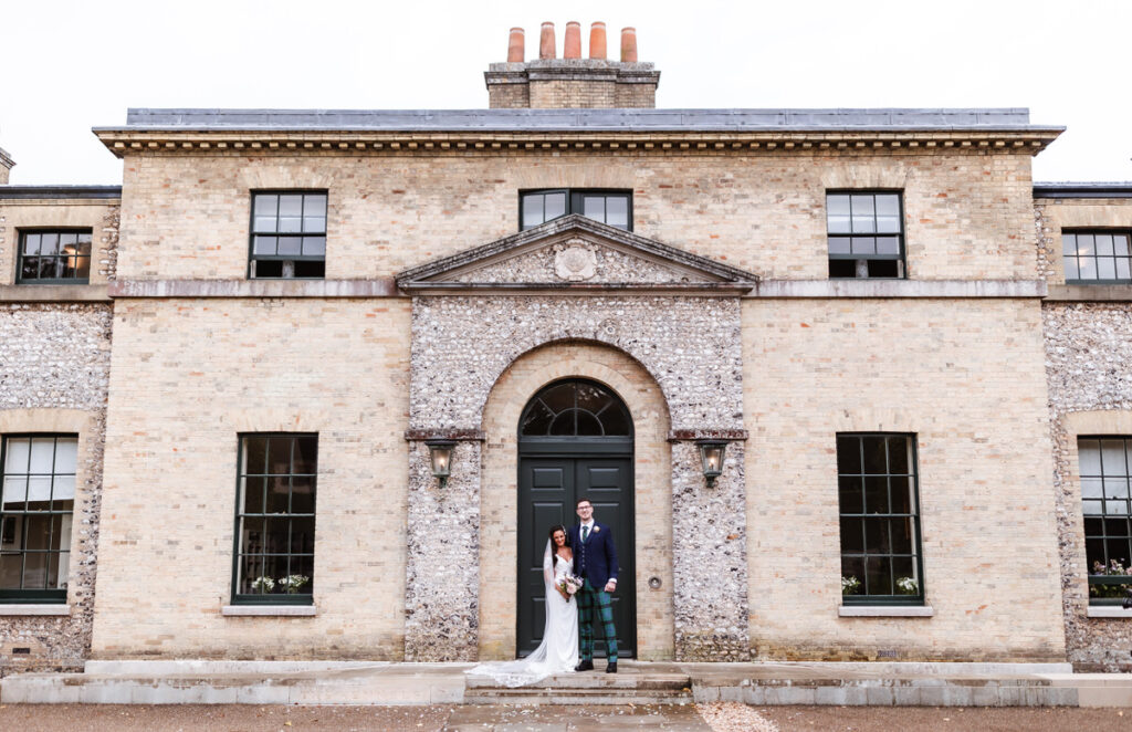 A bride and groom stand together in wedding attire on the steps in front of a large, beige brick building with tall windows and a central arched doorway.