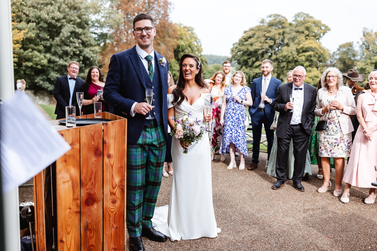 A bride in a white dress and a groom in a navy jacket and green tartan trousers stand smiling outdoors, surrounded by well-dressed guests at a wedding reception. Trees and greenery are visible in the background.