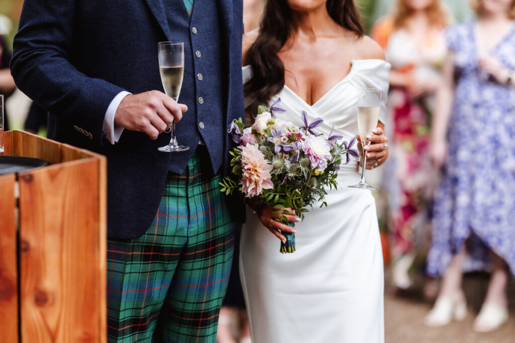 A bride in an off-shoulder white dress holds a bouquet and a champagne glass, standing beside a groom in a dark jacket and colorful tartan trousers, with guests blurred in the background.