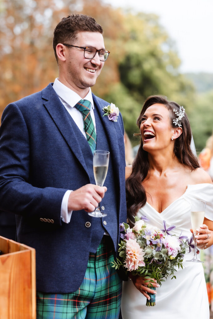 A smiling bride in a white dress holding a bouquet stands next to a groom in a blue jacket and green tartan, both holding champagne glasses and laughing outdoors.