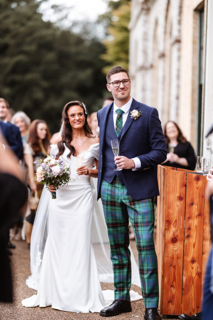A bride in a white dress and a groom in a navy jacket and green tartan pants stand smiling outdoors, with the bride holding a bouquet and people gathered around them.