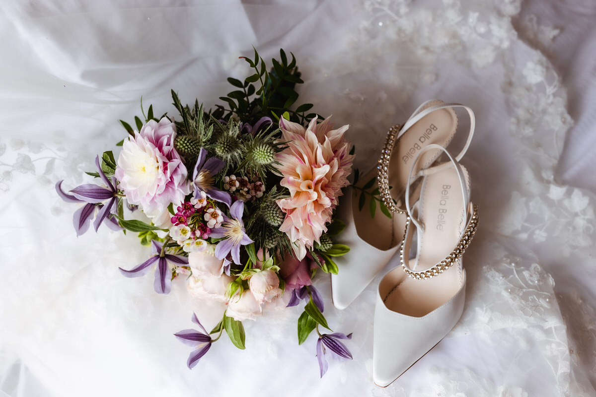 A bouquet of pastel flowers with greenery lies next to a pair of elegant white bridal shoes with jeweled straps, all arranged on delicate white lace fabric.