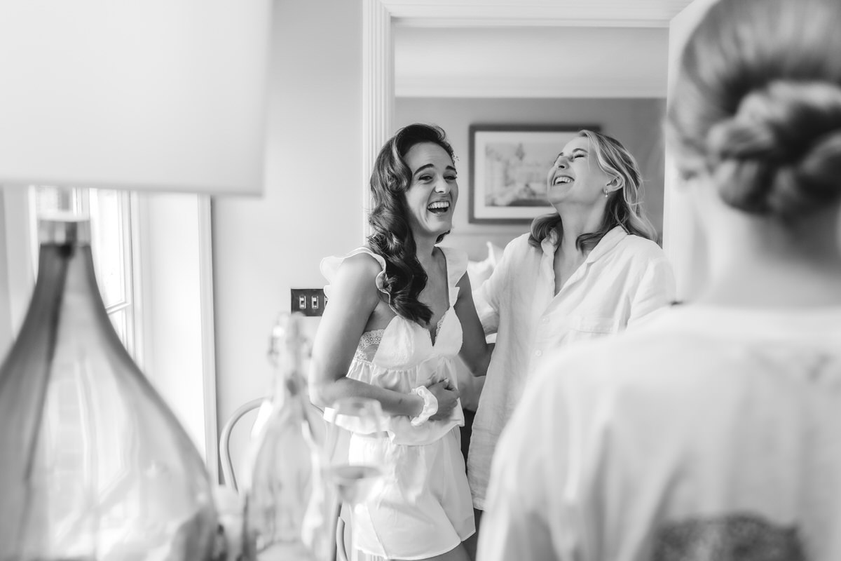 Two women stand near a window, laughing and smiling, while another person with hair in a bun faces them in the foreground. The scene is bright and joyful, captured in black and white.