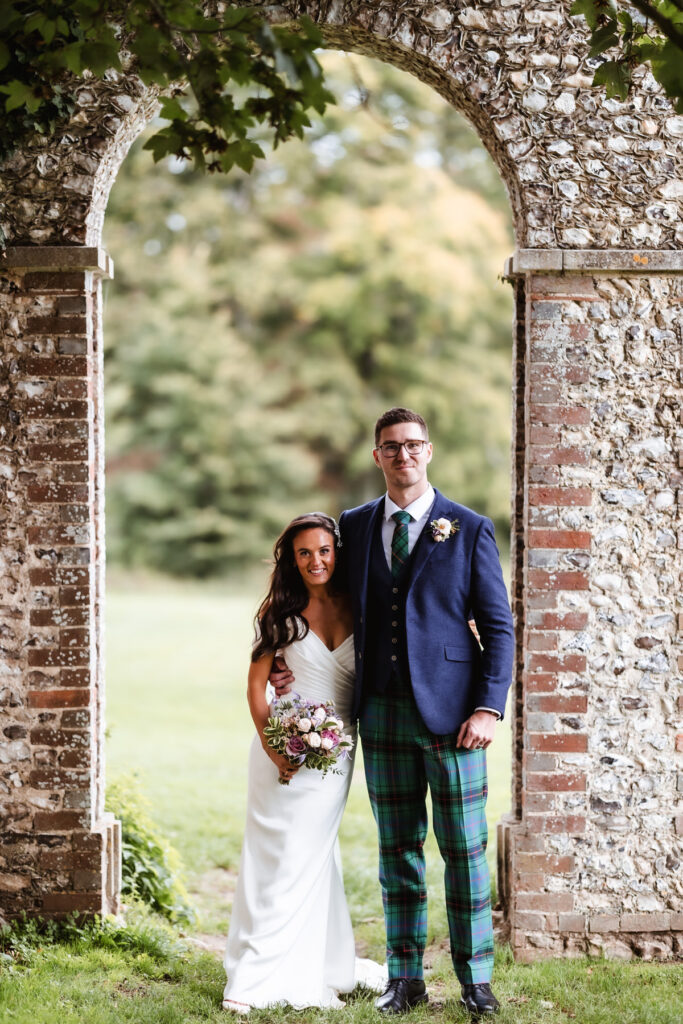 A bride in a white dress holding a bouquet stands beside a groom in a blue jacket and green tartan trousers, both smiling under a stone archway with greenery in the background.