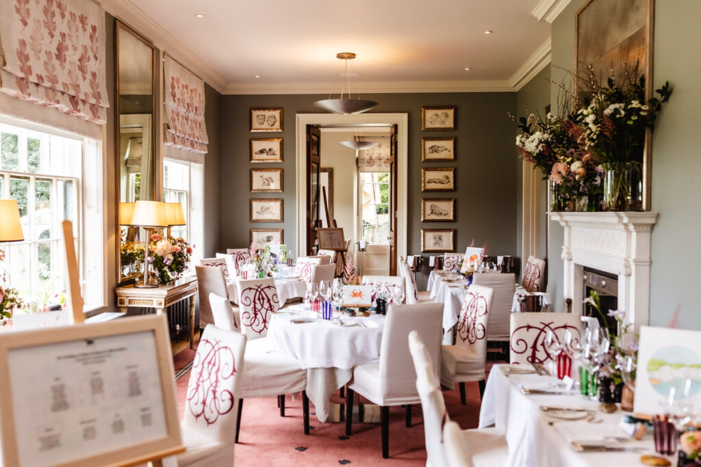 A bright, elegant dining room with round tables covered in white cloths, set for a meal. Large windows, floral arrangements, framed artwork, and soft lighting create a sophisticated, inviting atmosphere.