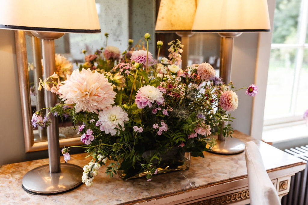 A floral arrangement with pink and white dahlias and greenery sits on a marble-topped table between two lamps, in front of a mirror in a softly lit room.