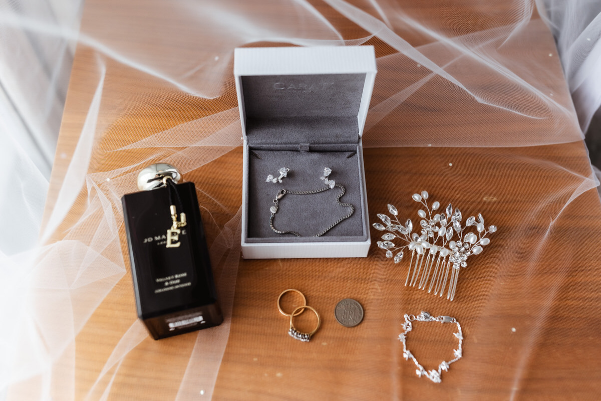 A bottle of Jo Malone perfume, two gold rings, a silver coin, a silver bracelet, a jeweled hair comb, and a jewelry box with a silver necklace and earrings are arranged on a wooden surface with sheer white fabric.