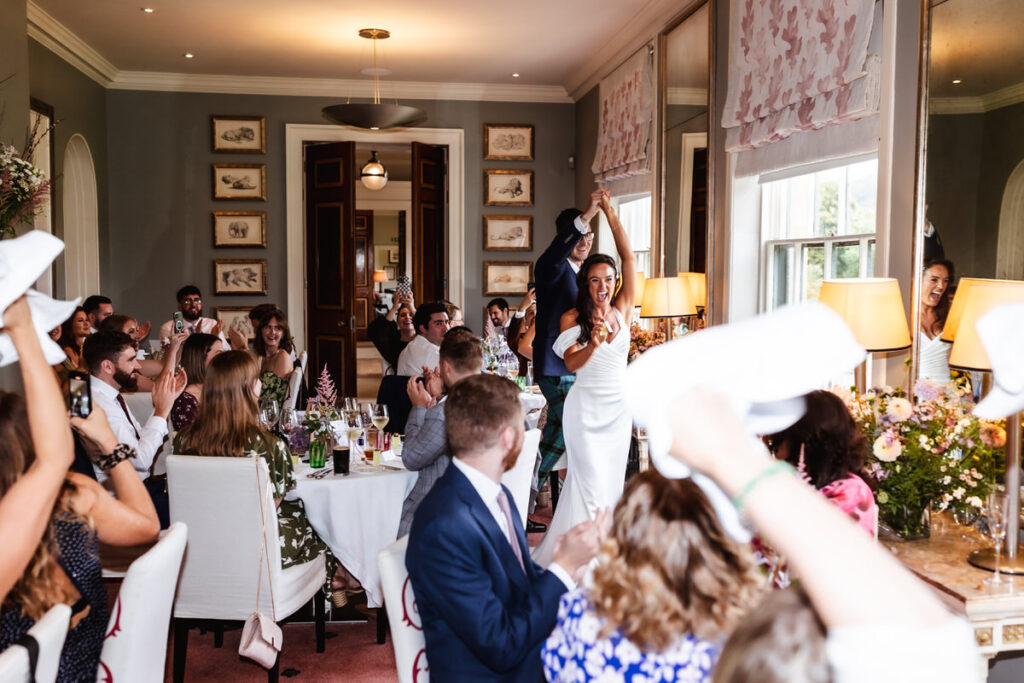 A bride and groom enter a bright, elegant dining room as guests cheer and wave napkins. The bride smiles, raising her bouquet, while the groom holds her hand, and sunlight streams through large windows.