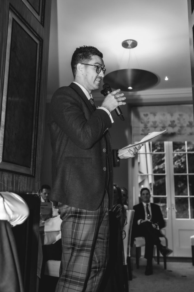 A man wearing glasses and plaid trousers speaks into a microphone while holding papers, standing near a doorway in a formal room with seated guests in the background. The image is in black and white.