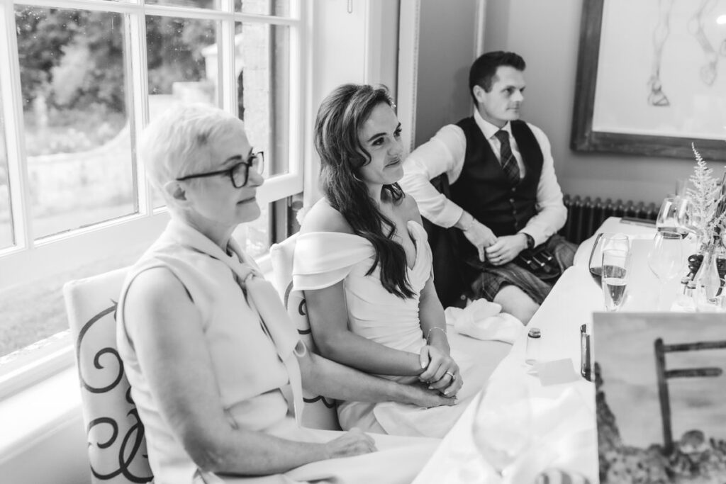 A black and white photo of two women and a man sitting at a table by a window. The woman in the center wears a wedding dress, while the other woman wears glasses. The man wears a vest and kilt. They appear to be listening intently.