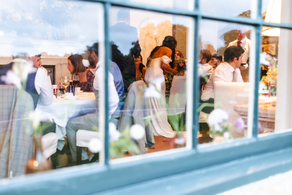 A wedding reception seen through a window, with guests seated at decorated tables, eating and talking. A bride in a white dress hugs a guest, while others enjoy the celebration indoors.
