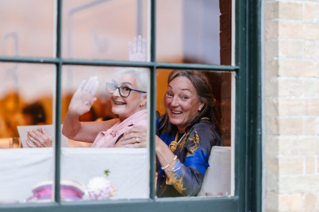 Two older women sit inside by a window, smiling and waving at the camera. One wears glasses and a pink scarf, the other wears a patterned blue top. The scene is cheerful and friendly.