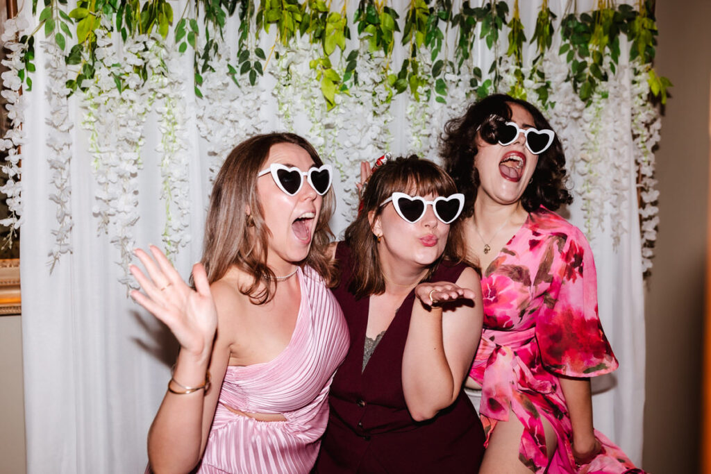 Three women in party outfits and heart-shaped sunglasses pose playfully in front of a white curtain decorated with greenery and flowers, smiling, waving, and blowing a kiss at the camera.
