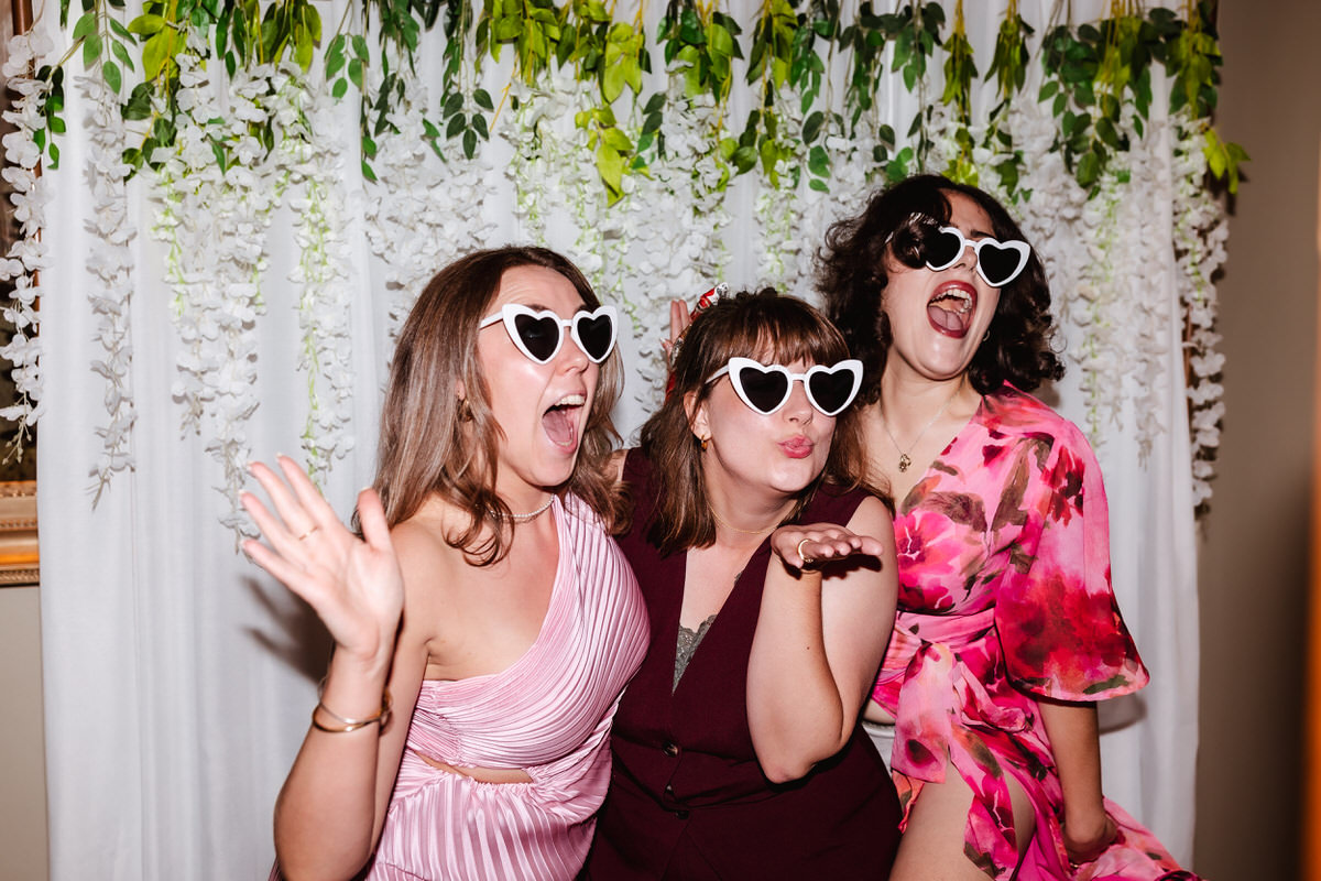 Three women in party outfits and heart-shaped sunglasses pose playfully in front of a white curtain decorated with greenery and flowers, smiling, waving, and blowing a kiss at the camera.