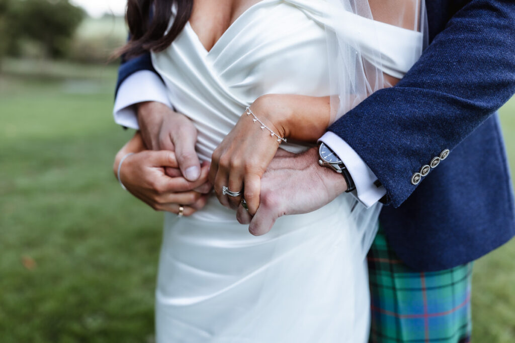 A close-up of a bride and groom embracing outdoors, with the groom’s arms wrapped around the bride’s waist. The bride wears a white dress and jewelry; the groom wears a blue jacket and tartan kilt. Green grass is visible in the background.