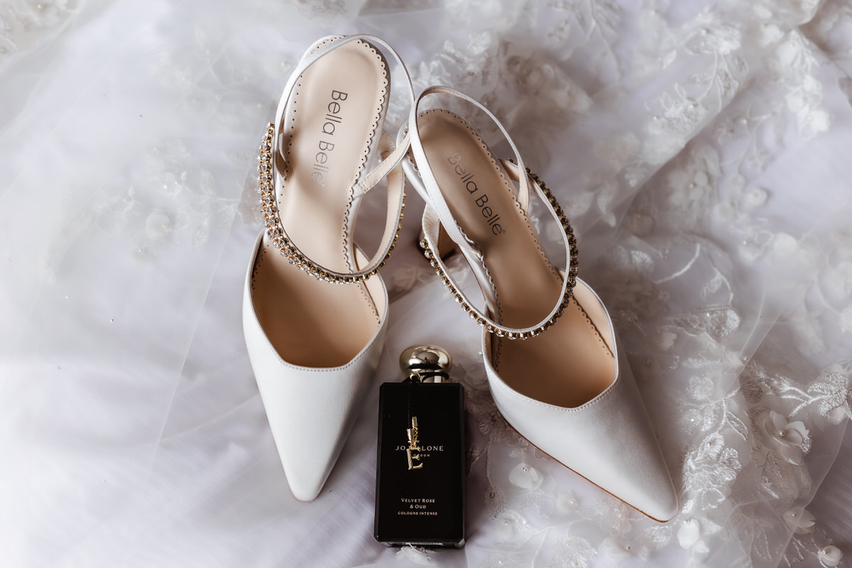 A pair of white Bella Belle high-heeled shoes with jeweled ankle straps sit on a lacy white fabric next to a black Yves Saint Laurent Libre perfume bottle.