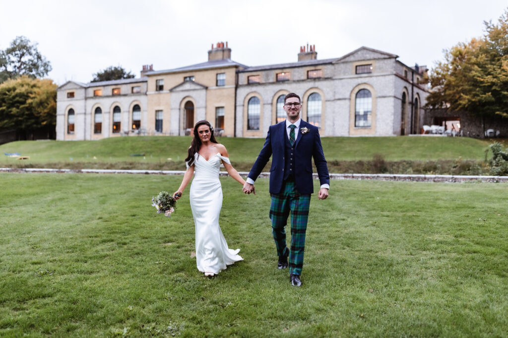 A bride in a white dress and a groom in a navy jacket and green tartan trousers walk hand in hand on a grassy lawn in front of a large, historic building.