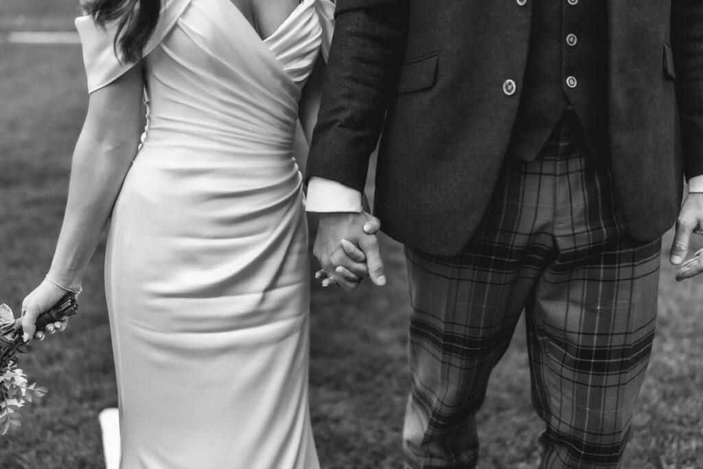 Black and white photo of a couple holding hands outdoors. The woman wears a draped, off-shoulder gown and holds a bouquet; the man wears a suit jacket and plaid trousers. Only their bodies are visible, not their faces.