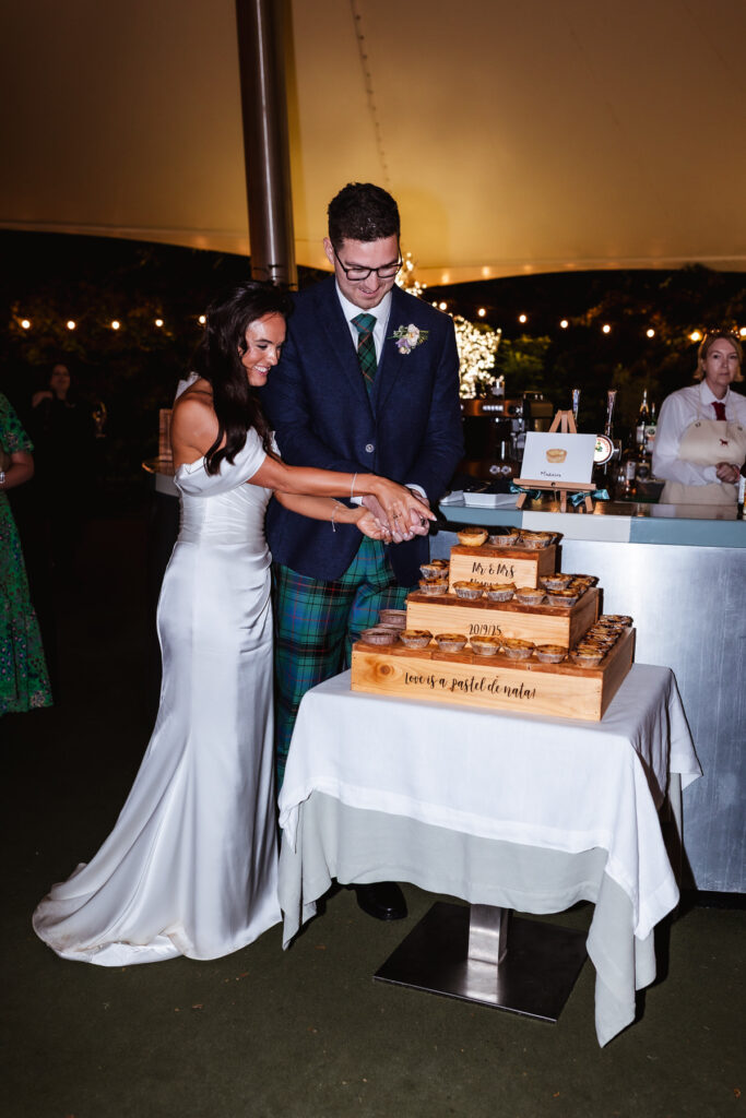 A bride in a white dress and groom in a dark jacket and tartan pants smile as they cut a rustic wooden wedding cake together. The cake sits on a table in front of them, surrounded by guests and bar staff.