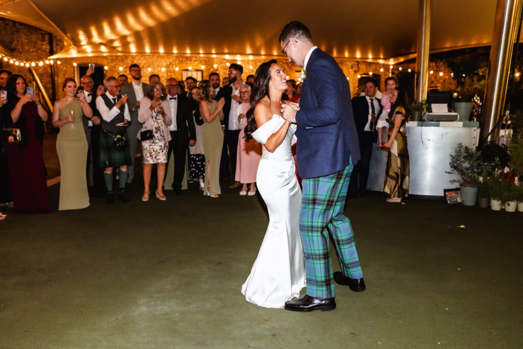 A bride and groom share their first dance under a tent, surrounded by guests in formal attire, with warm string lights creating a festive atmosphere.